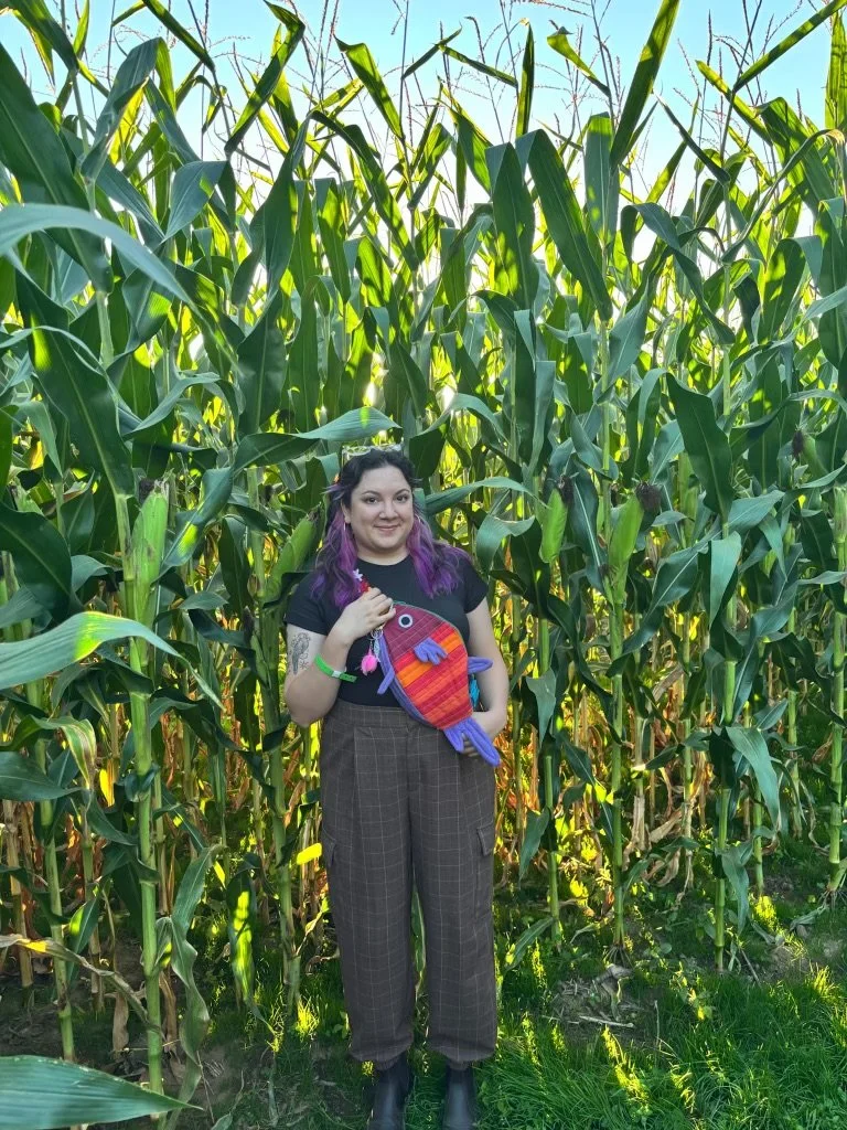 A woman with purple hair standing in a cornfield, holding a colorful fish-shaped stuffed toy.