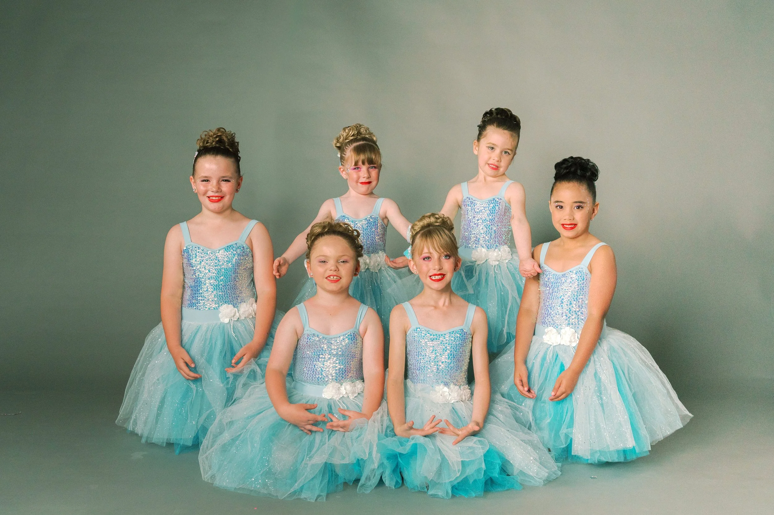 Seven young girls dressed in matching blue sequined ballet costumes with tulle skirts, posing together against a plain background.