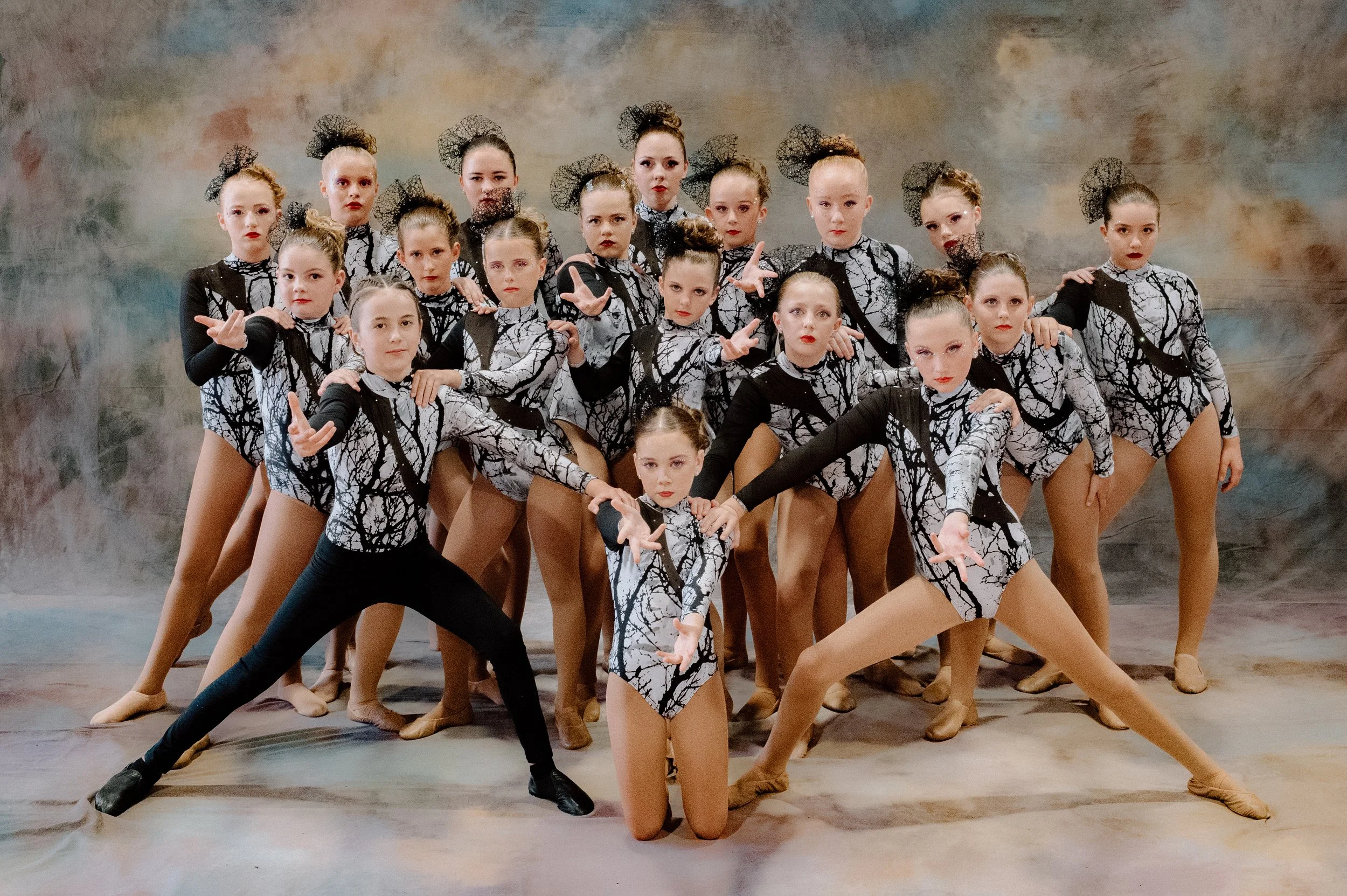 Group of young girls dressed in matching purple and black costumes, posing together with some kneeling in front and others standing behind them, arms raised and smiling, against a neutral background.