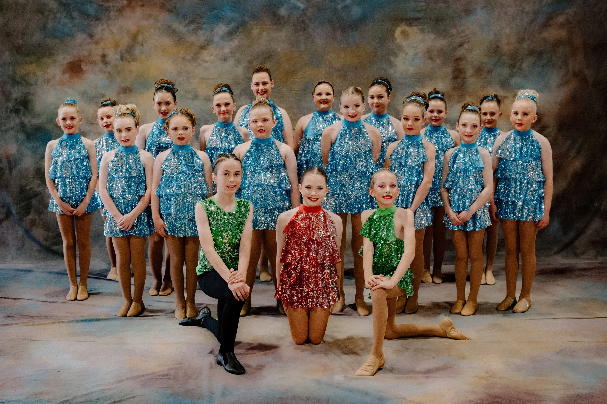 A group of young female dancers in shiny silver costumes, posing for a photo with some standing and some kneeling, against a gray background.