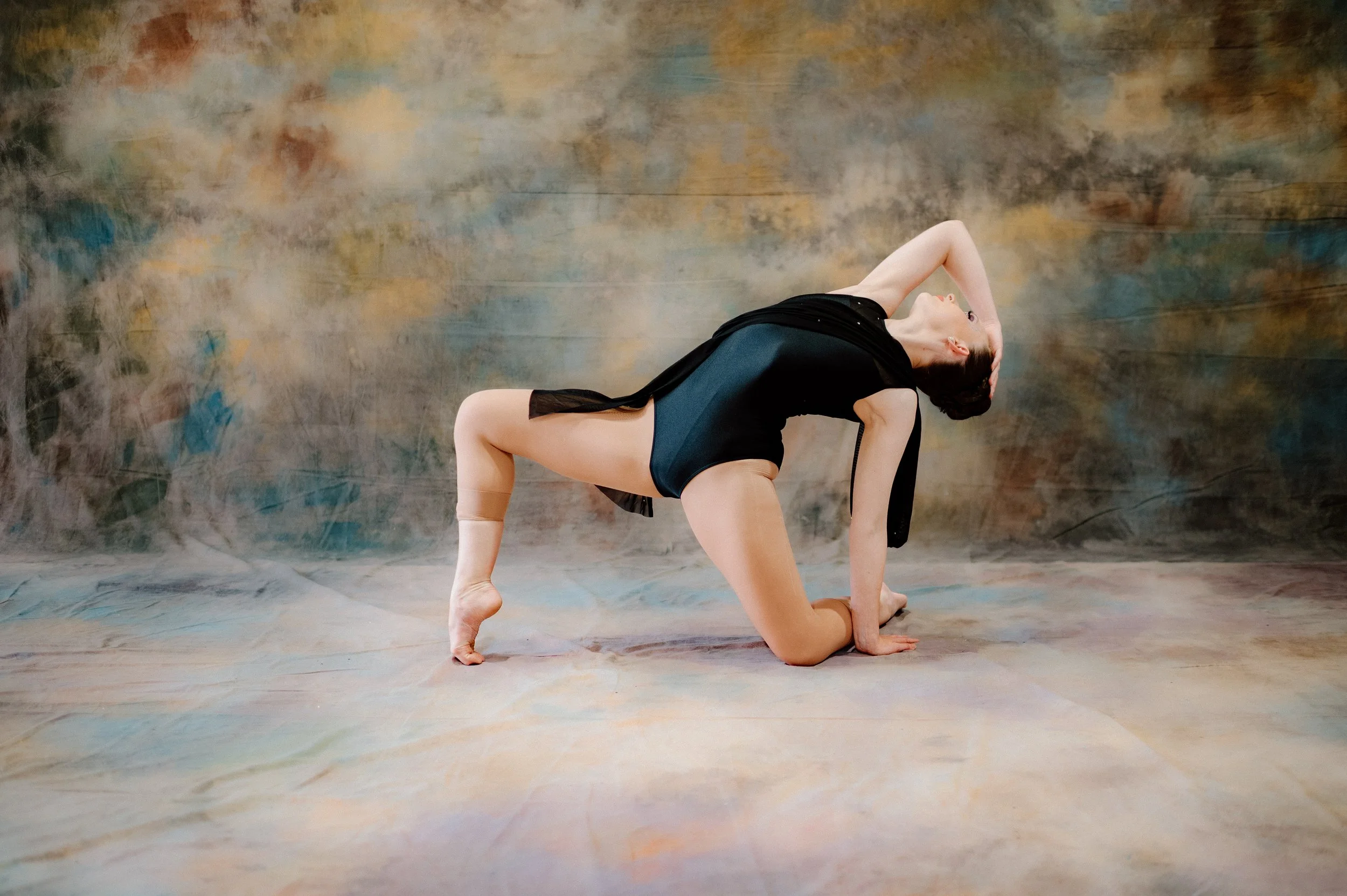 Young girl performing a dance move on a gray floor, wearing a pink and white varsity jacket, gray shirt, black Adidas track pants, and black shoes, with braided hair and looking at the camera.