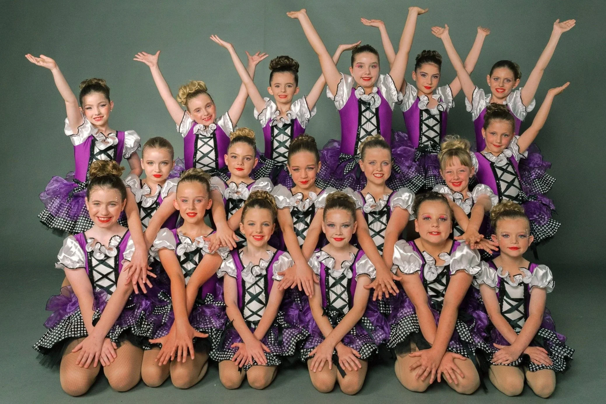 Group of young girls dressed in matching purple and black costumes, posing together with some kneeling in front and others standing behind them, arms raised and smiling, against a neutral background.