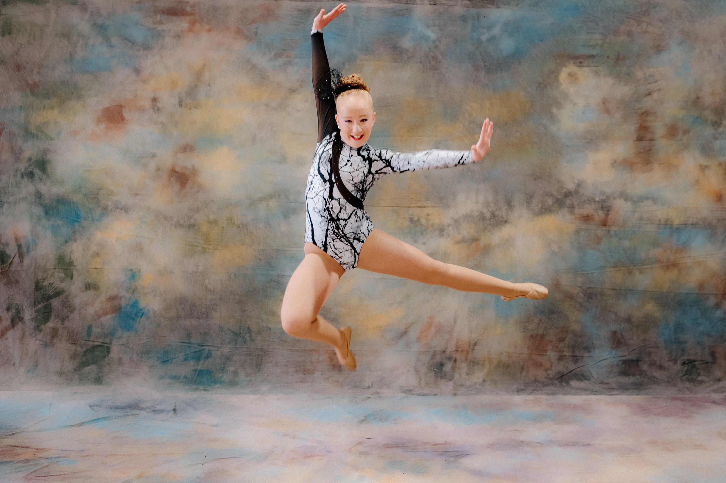 Young female figure skater in a silver and blue costume, performing a pose with arms outstretched against a plain background.