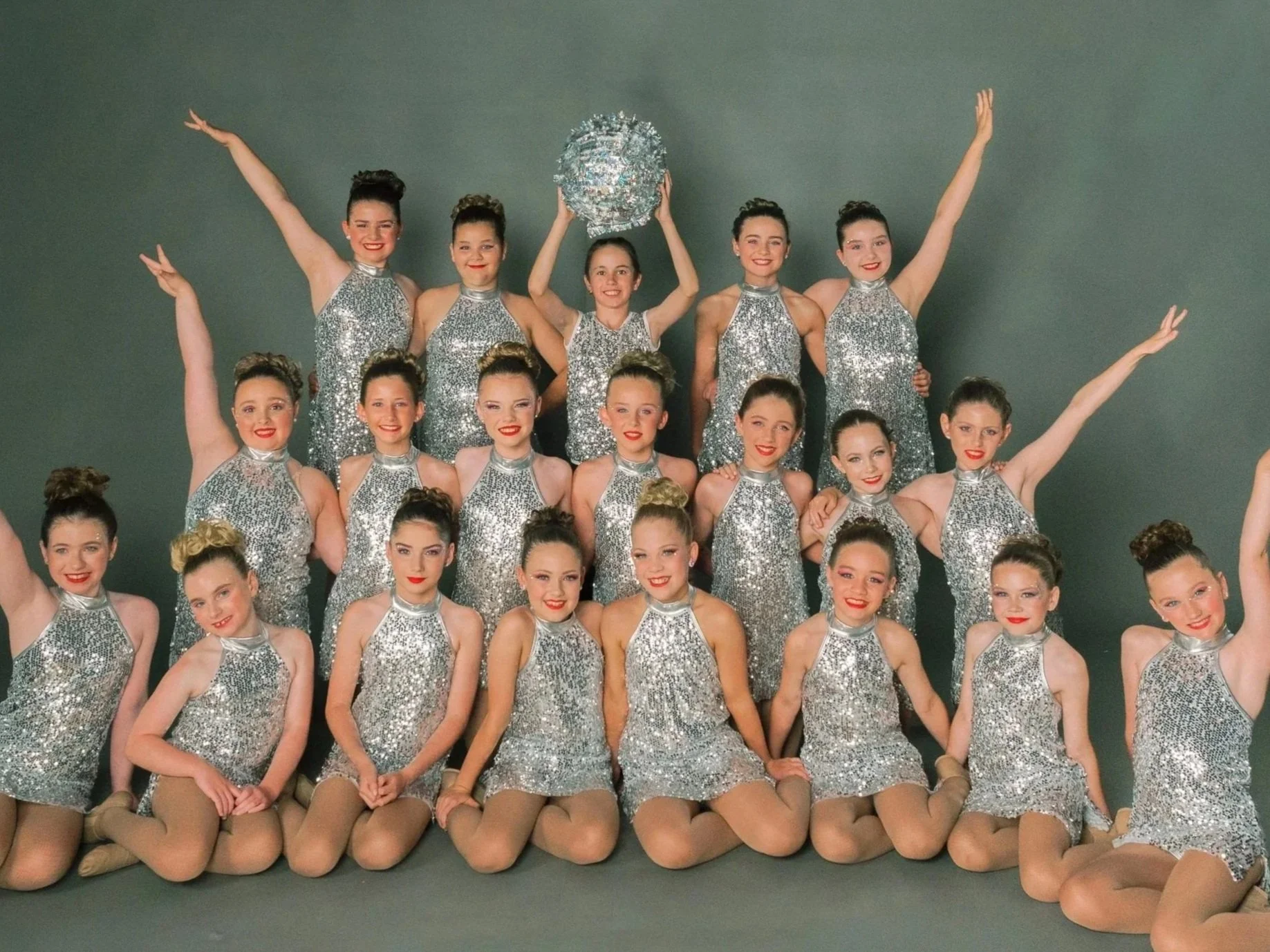 A group of young female dancers in shiny silver costumes, posing for a photo with some standing and some kneeling, against a gray background.