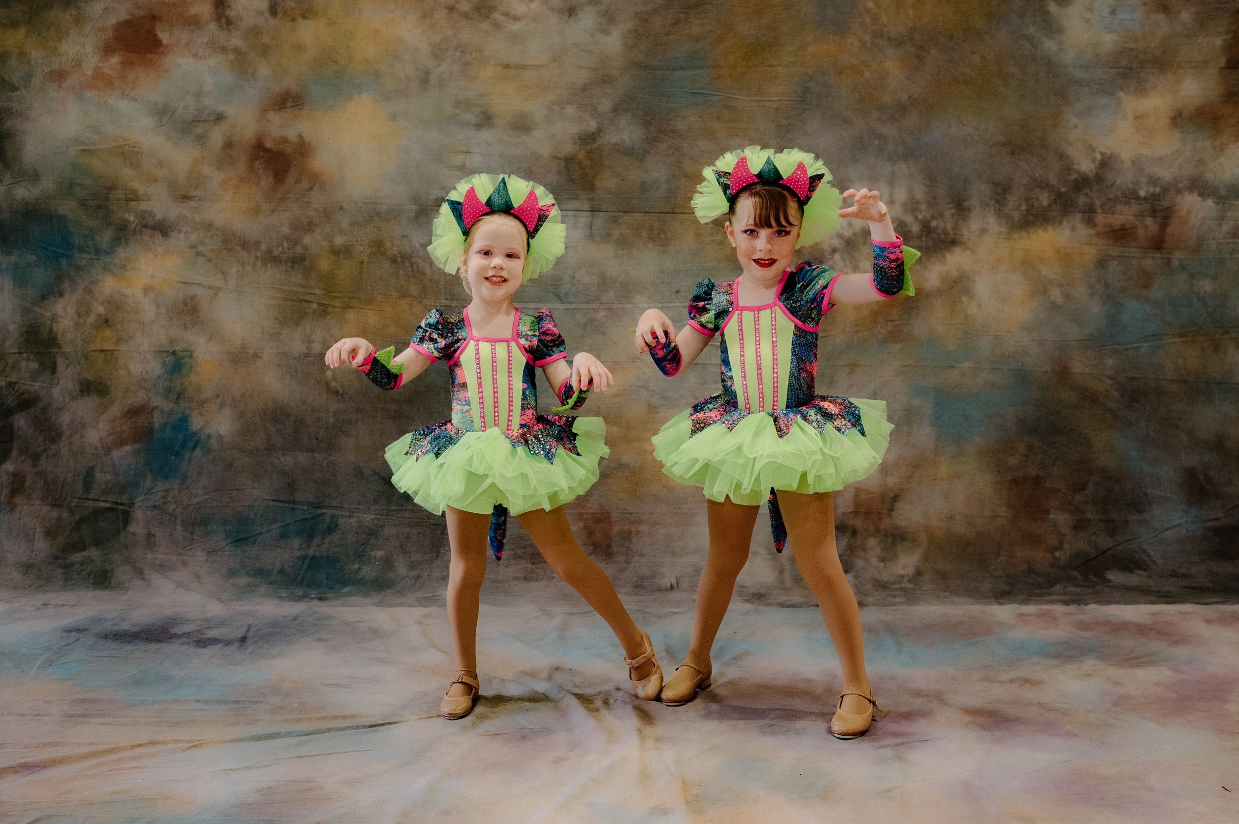 A young girl with a pink bow in her hair, lying on her stomach on a gray surface, resting her chin on her hands, smiling at the camera, wearing a colorful ruffled outfit.