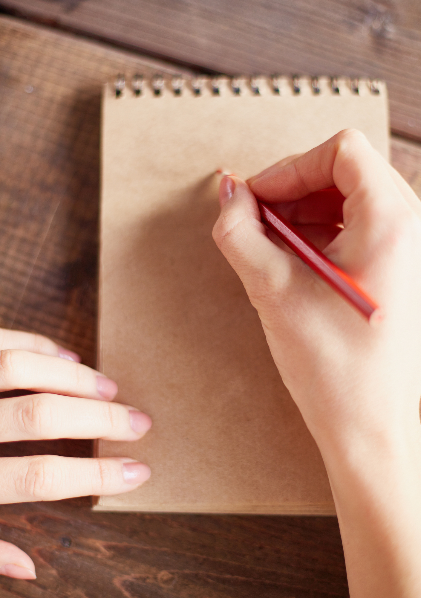 A person writes on a blank spiral notebook with a red pencil. Hands are focused, suggesting creativity. The wooden table background adds warmth.