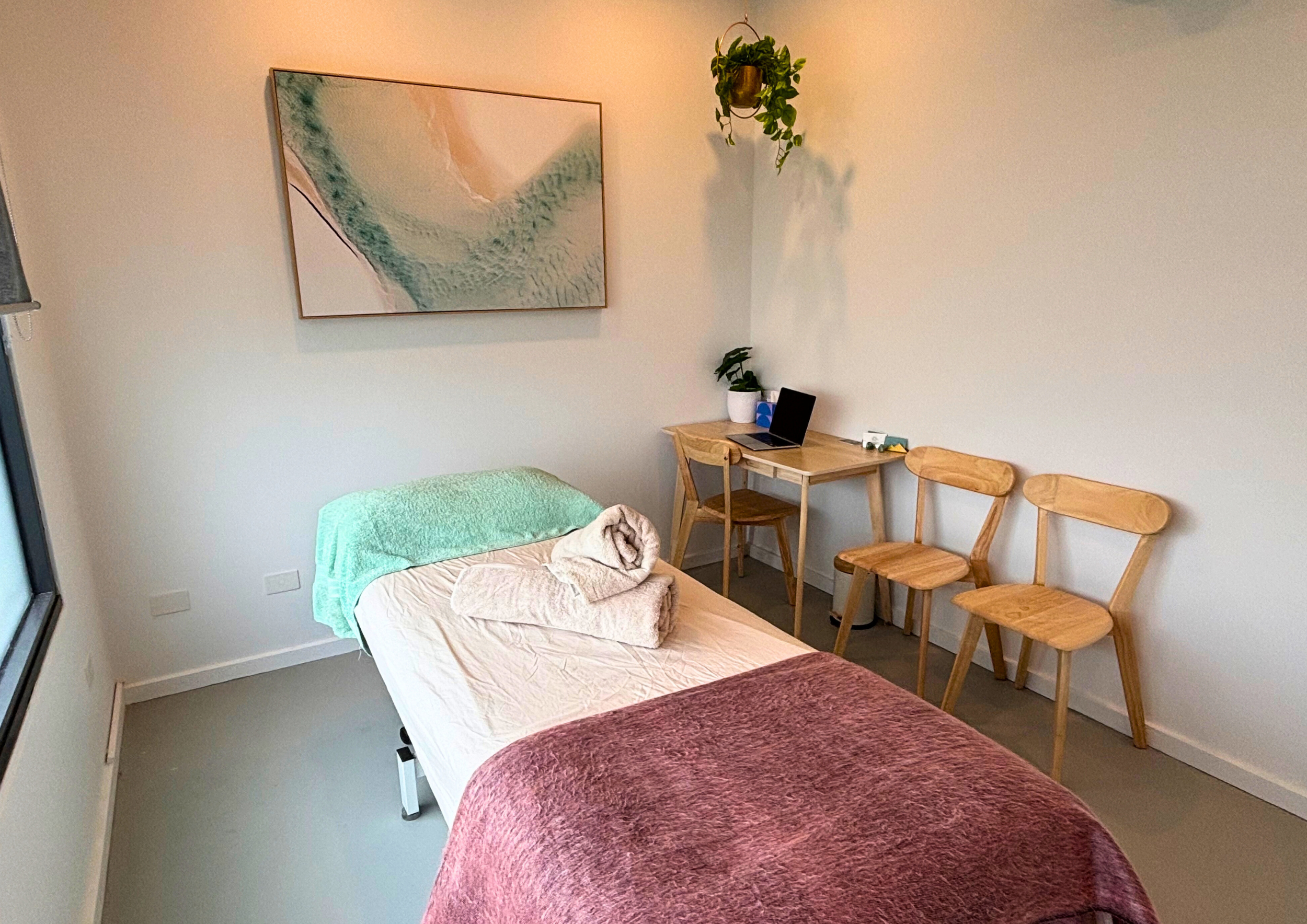 A bedroom with two beds, one with a pinkish-burgundy cover and the other with a beige cover with a green towel on it, against a light-colored wall. On the wall, there is a large abstract painting with pastel colors. To the right, there is a small wooden desk with a laptop, potted plants, and some items, and next to it three wooden chairs. Above the desk, a hanging plant casts shadows on the wall.