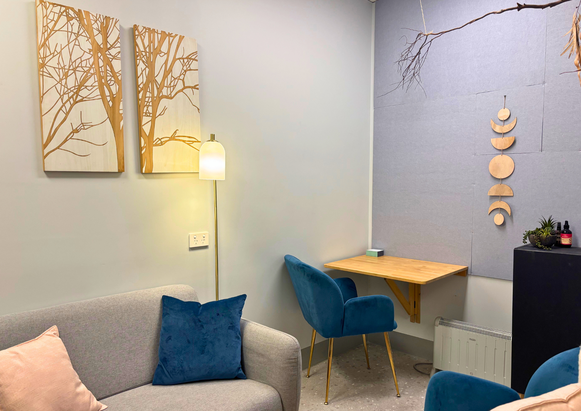 Cozy room with grey walls, blue chairs, and a grey couch. Wooden tree artwork and geometric wall decor add warmth. A small wooden desk fits the serene space.