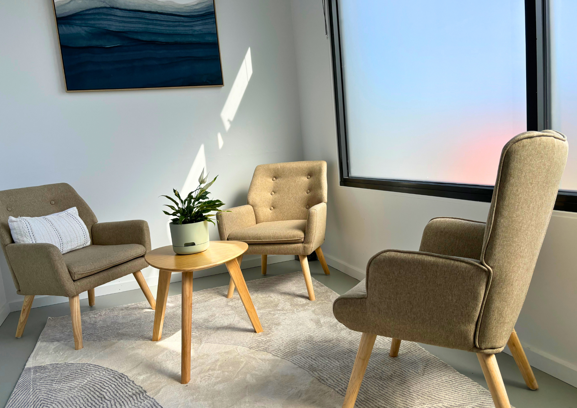 Cozy room with three beige armchairs around a round wooden table. A potted plant sits on the table. A large abstract painting and soft sunlight create a calm atmosphere.