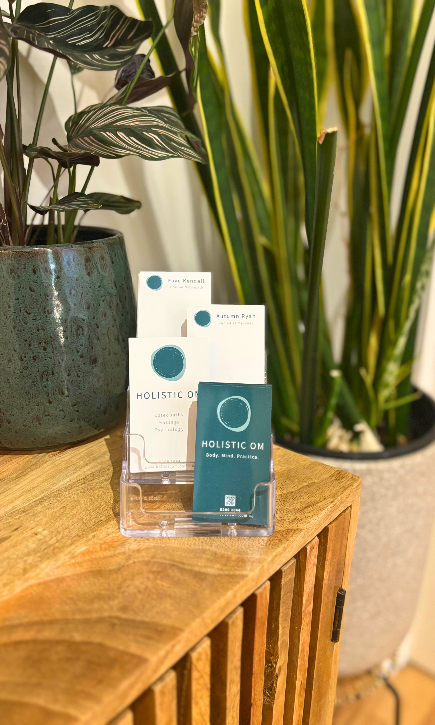 Holistic OM Business Cards stating Osteopathy, Massage Psychology and Autumn Ryan and Faye Kendall on a wooden bench surrounded by plants