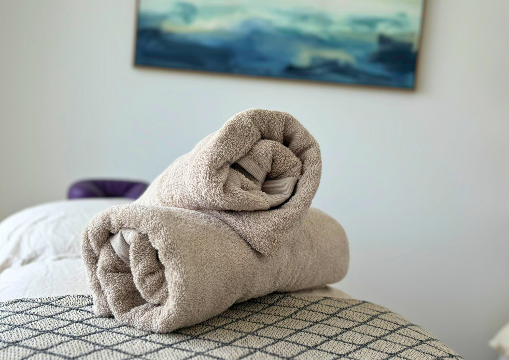 Two neatly rolled beige towels on a patterned bedspread, with a blurred ocean-themed painting in the background, conveying a calm, tidy atmosphere.
