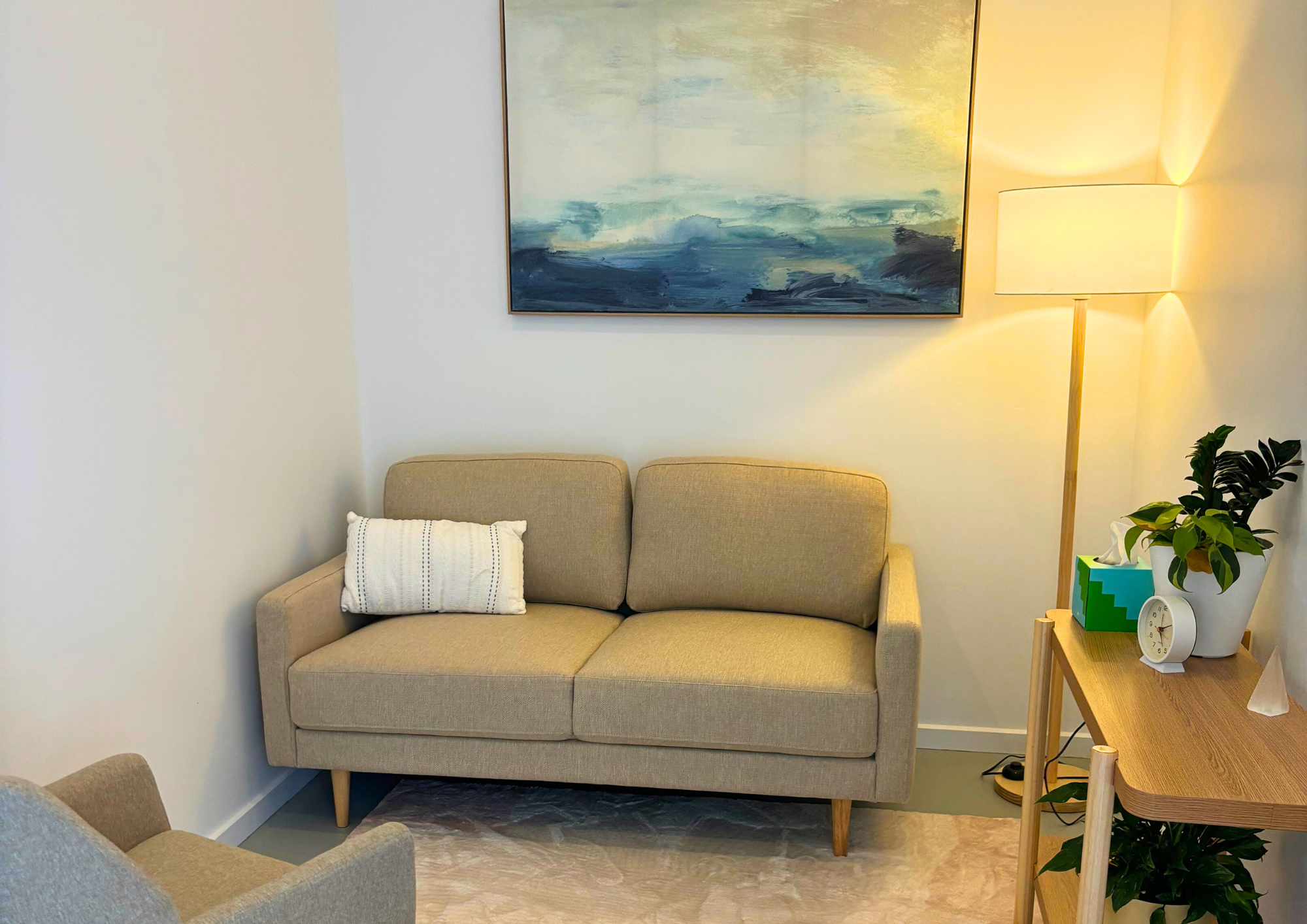 A cozy living room with a beige sofa and white pillow under a calming abstract ocean painting. A lit floor lamp and plants add warmth and serenity.