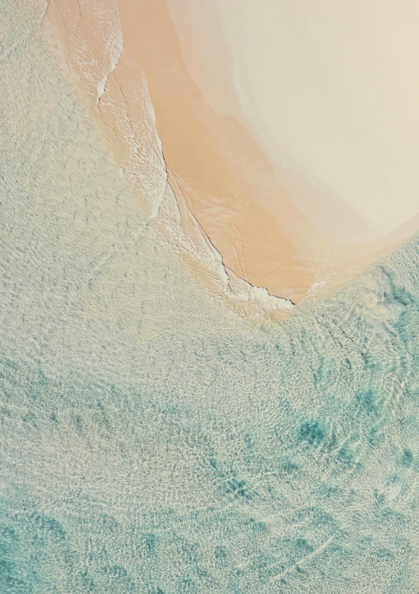 Aerial view of a tranquil beach showing soft beige sand merging with gentle turquoise waves, creating a serene and relaxing atmosphere. Close up of image in Holistic Om treatment room