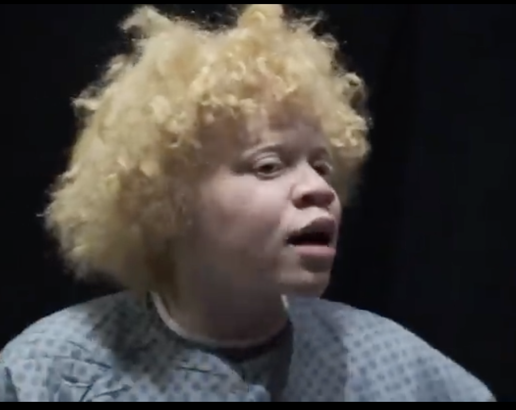 A woman in a hospital gown under stage lighting, mid-speech — Ava (MENTAL).