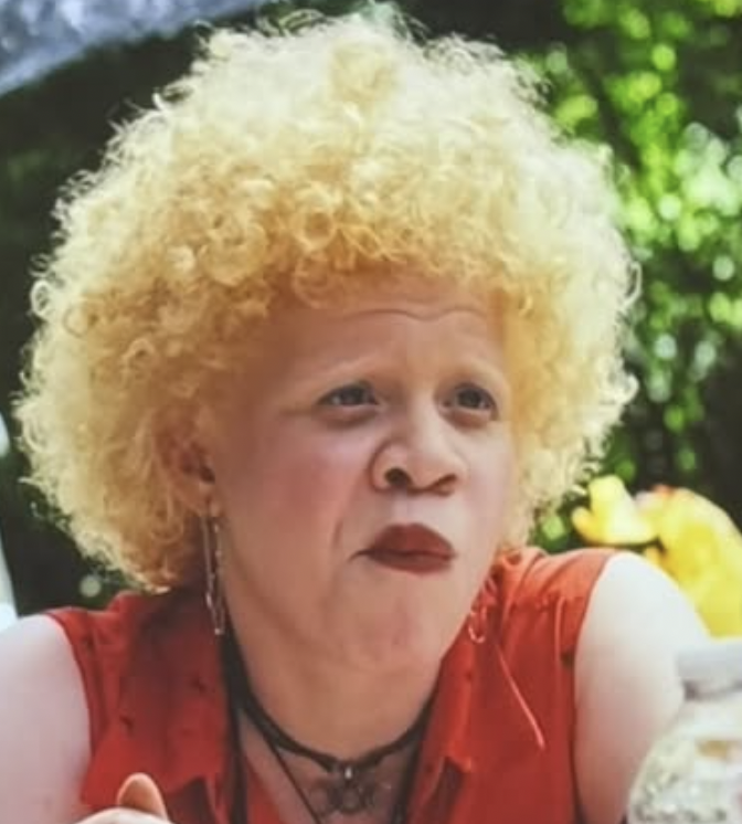 A woman in a sleeveless red top outdoors with curly blonde hair — Amber (Poker Face)