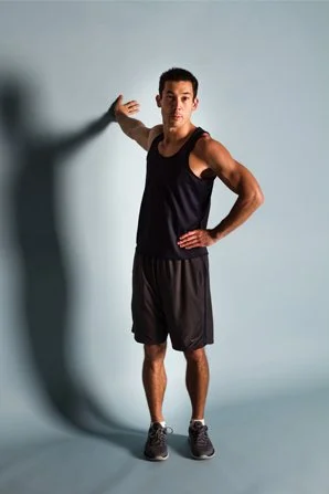 A man standing in a sports outfit with one hand on his hip and the other touching the wall, against a plain background.