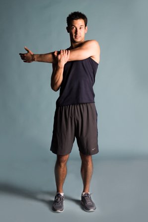 A man in athletic clothing stretching his arm across his chest in a gym or studio setting.