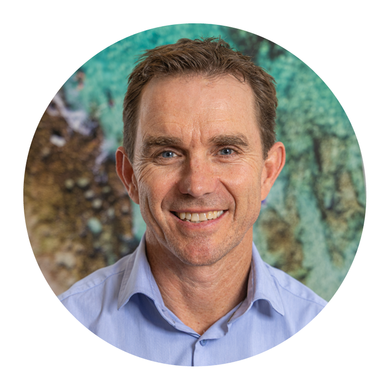 A middle-aged man with short brown hair, blue eyes, and a friendly smile, wearing a light blue collared shirt, standing in front of a colorful abstract background.