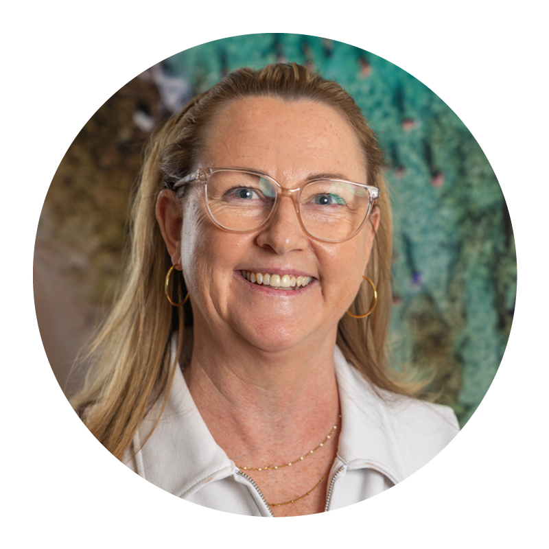 A smiling woman with blonde hair, glasses, and wearing gold hoop earrings and a white top with a zip-up collar, standing in front of a colorful, textured background.
