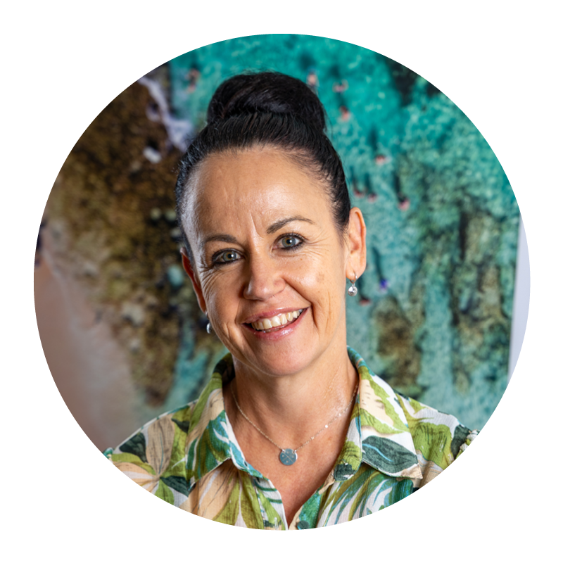 A woman with dark hair in a bun, smiling and wearing a green floral blouse, standing in front of an aquarium with fish and coral.