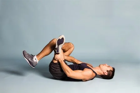 A woman lying on her back on a gray floor holding her knees to her chest in a stretch.