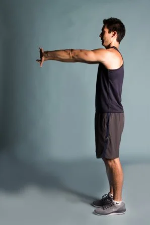 A man in workout clothes performing a front arm stretch against a plain light blue background.
