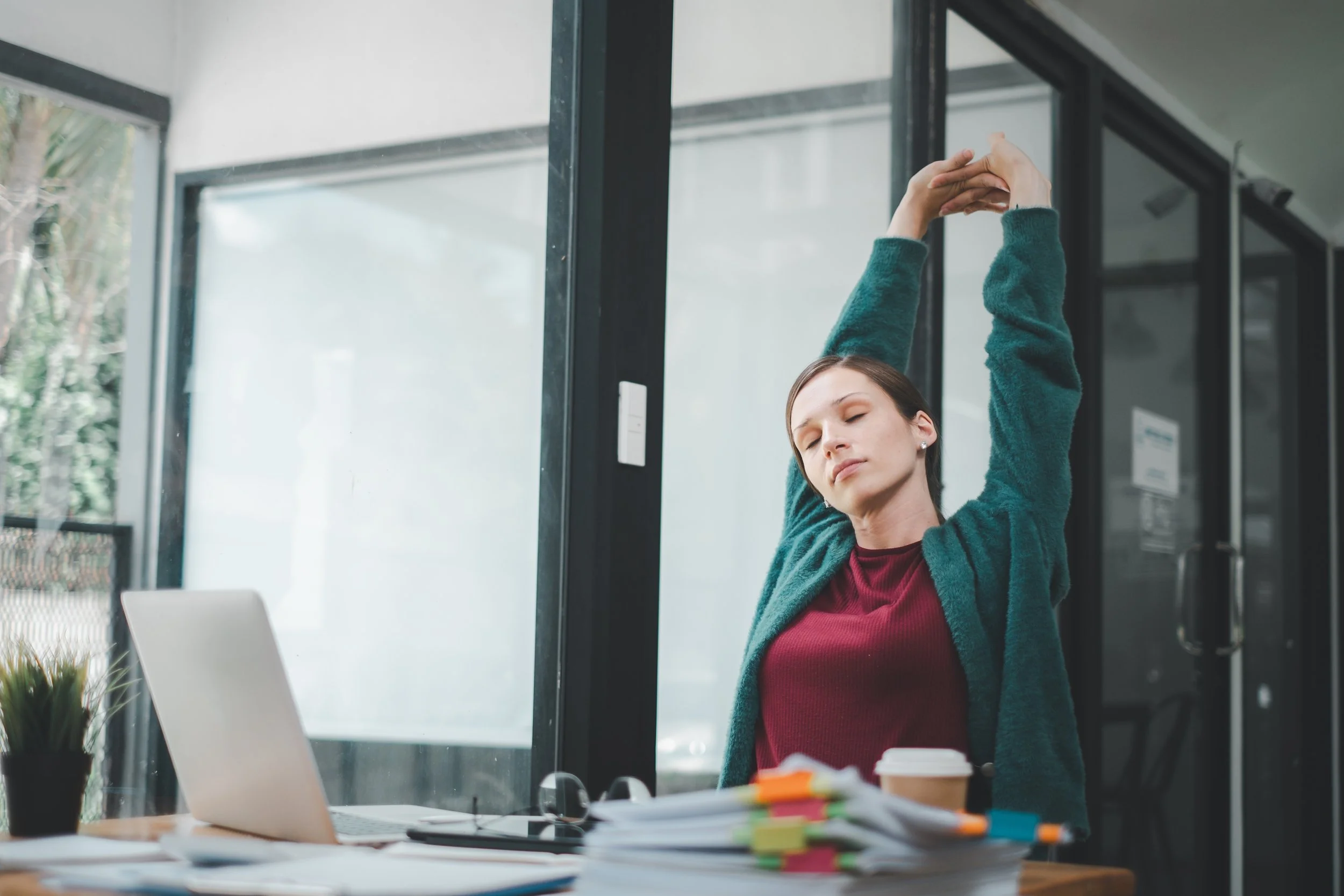 Young woman at her desk in an office, stretching with her arms raised above her head, surrounded by papers, a coffee cup, a potted plant, and an open laptop.