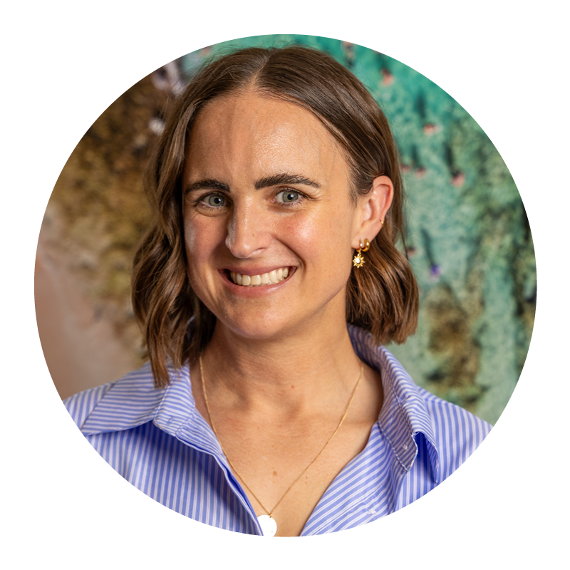 A woman with shoulder-length brown hair smiling while wearing a blue and white striped shirt, gold earrings, and a necklace, standing outdoors with trees and bushes in the background.