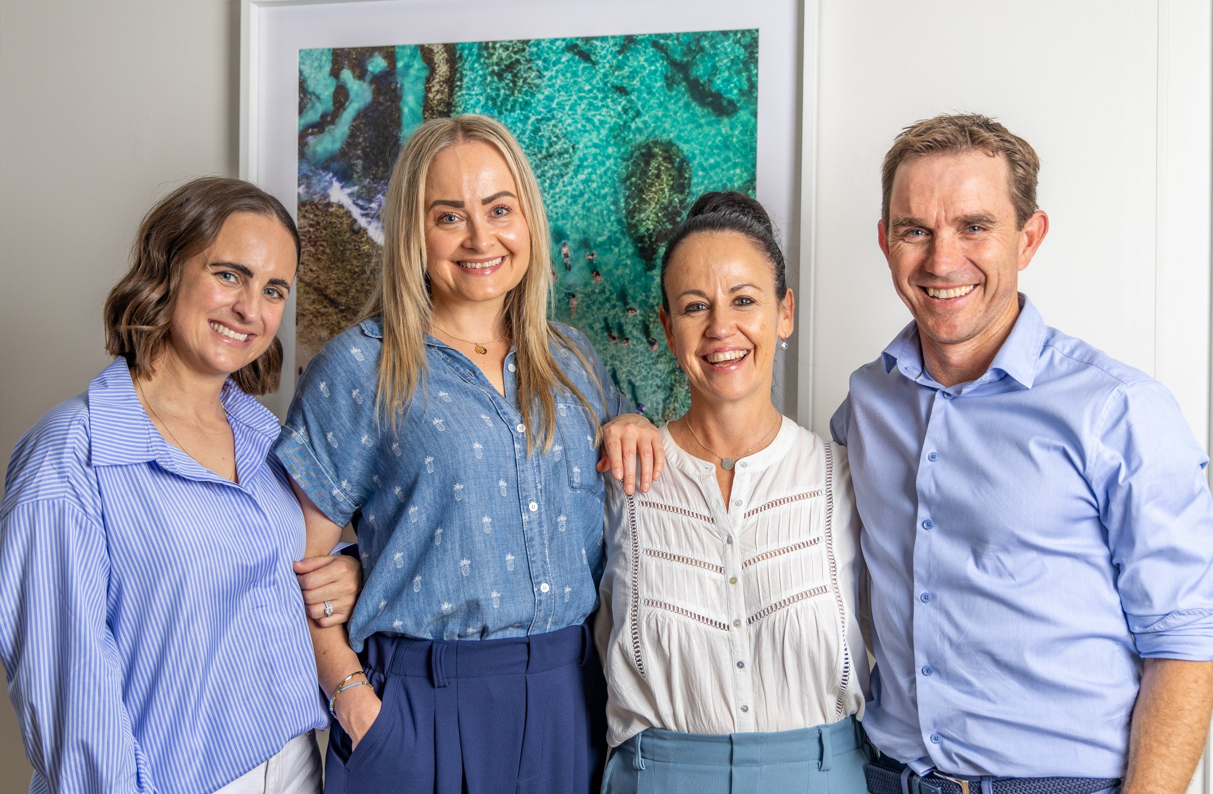 Four adults standing close together, smiling in front of a framed aerial view of a coastline with clear blue water.