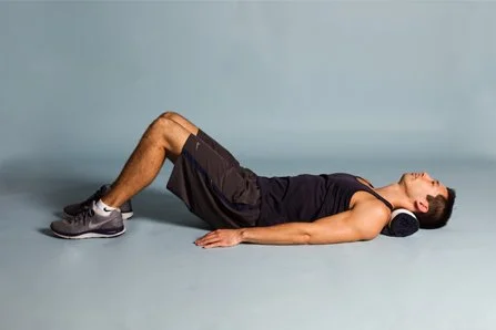A man lying on his back with a foam roller under his neck, wearing a black sleeveless shirt, black shorts, and sneakers, in a light-colored studio setting.