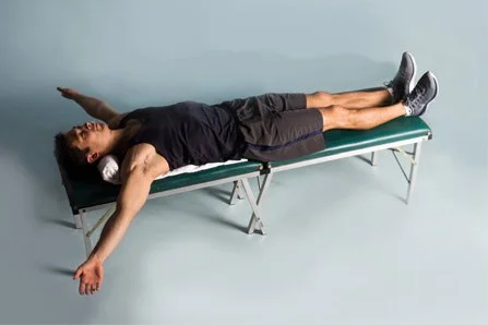 Person lying on a green massage table with arms and legs extended, eyes closed, in a relaxed position.