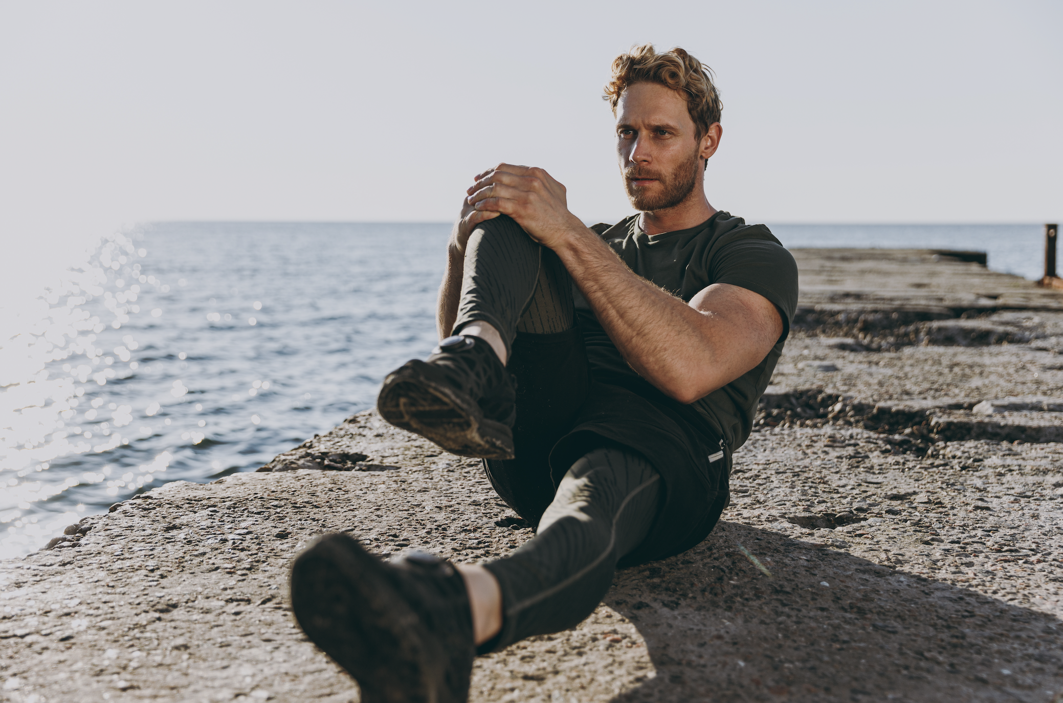 A man sitting on a concrete dock by the water holding his knee after exercising or stretching.