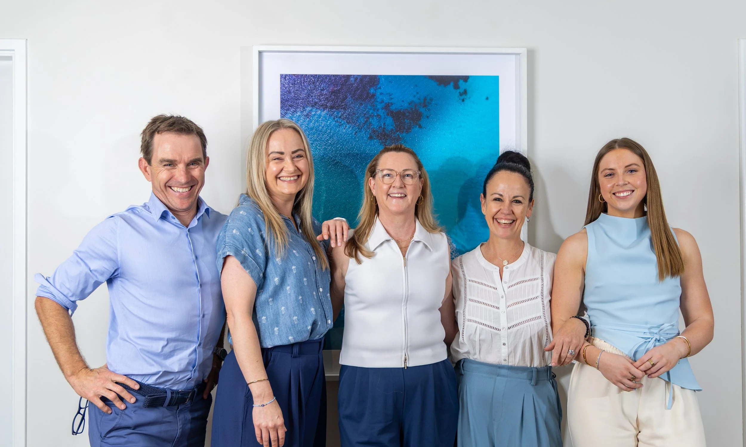 A group of five smiling professionals standing in front of abstract blue artwork in an office setting.