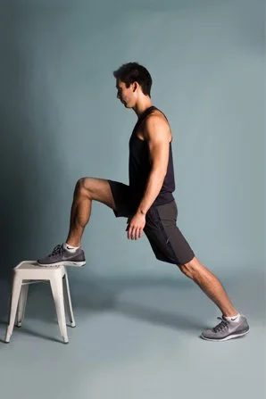 A man performing a stretch with one foot on a stool, lunging forward against a plain background.