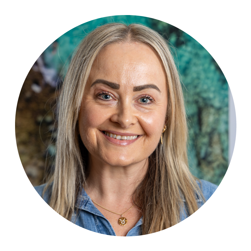 A smiling woman with blonde hair, wearing a denim shirt and a gold necklace, standing outdoors in front of a blurred nature background.