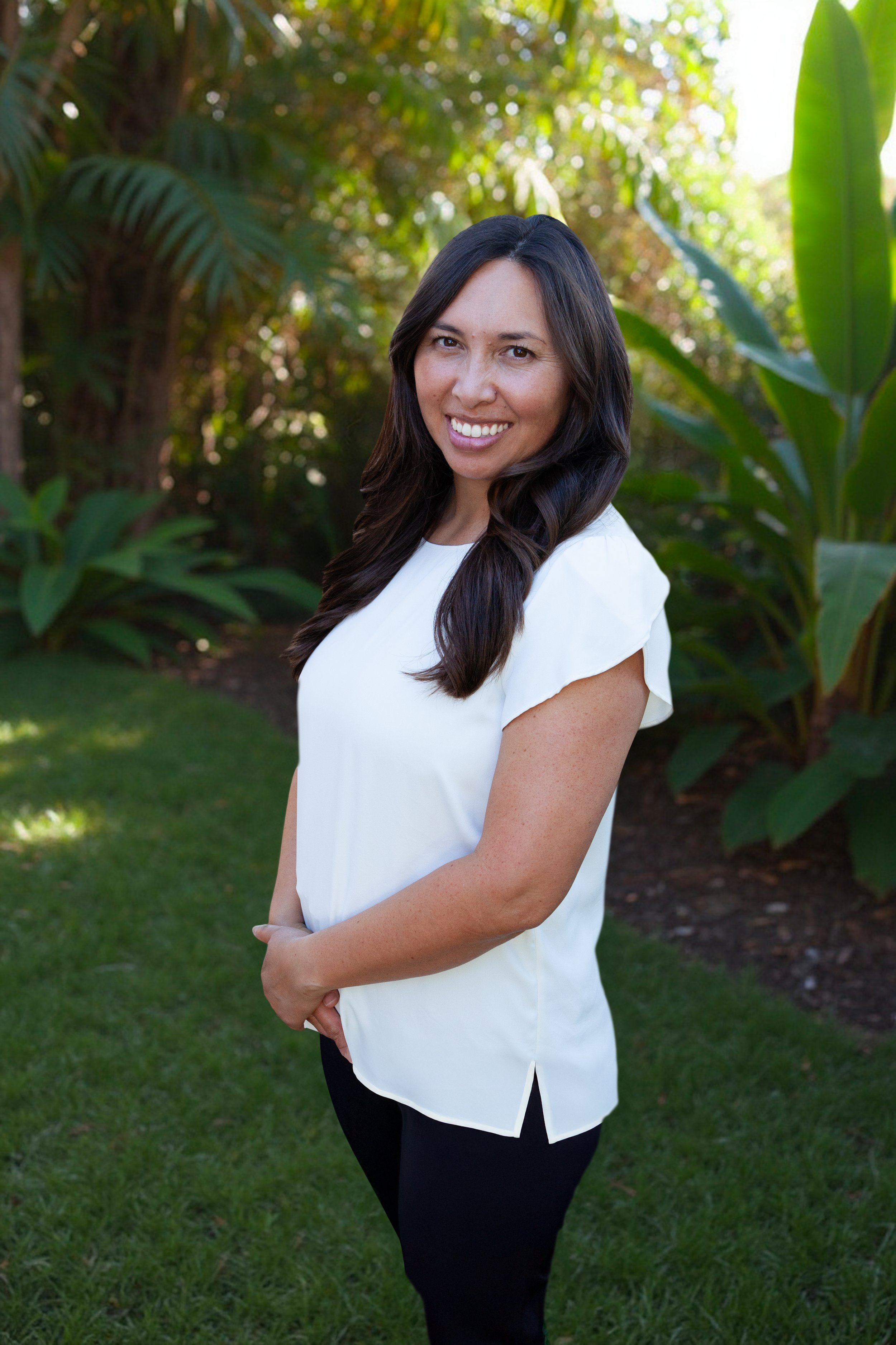 A smoking hot woman with long dark hair smiling outdoors, wearing a white blouse and black pants, standing in front of lush green plants.