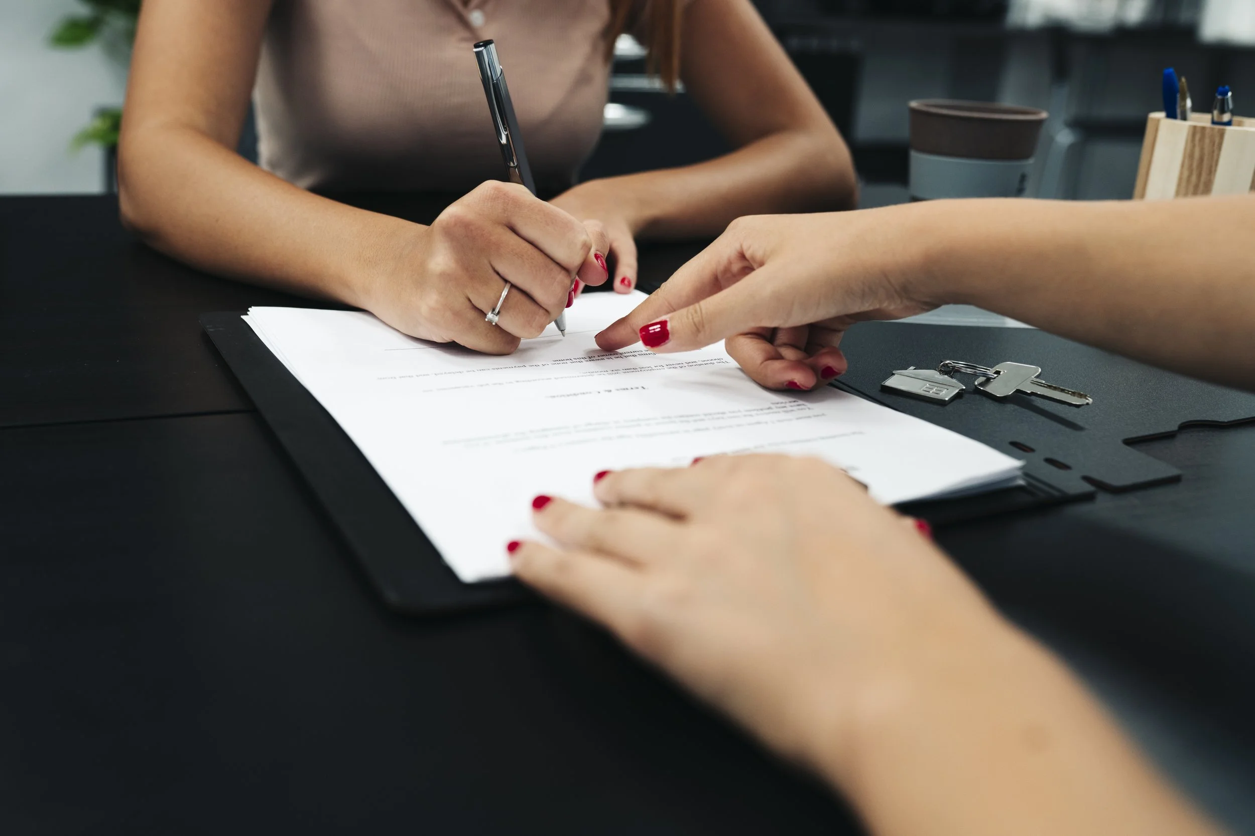 Two women are signing a notarized document at a desk with keys and office supplies nearby.