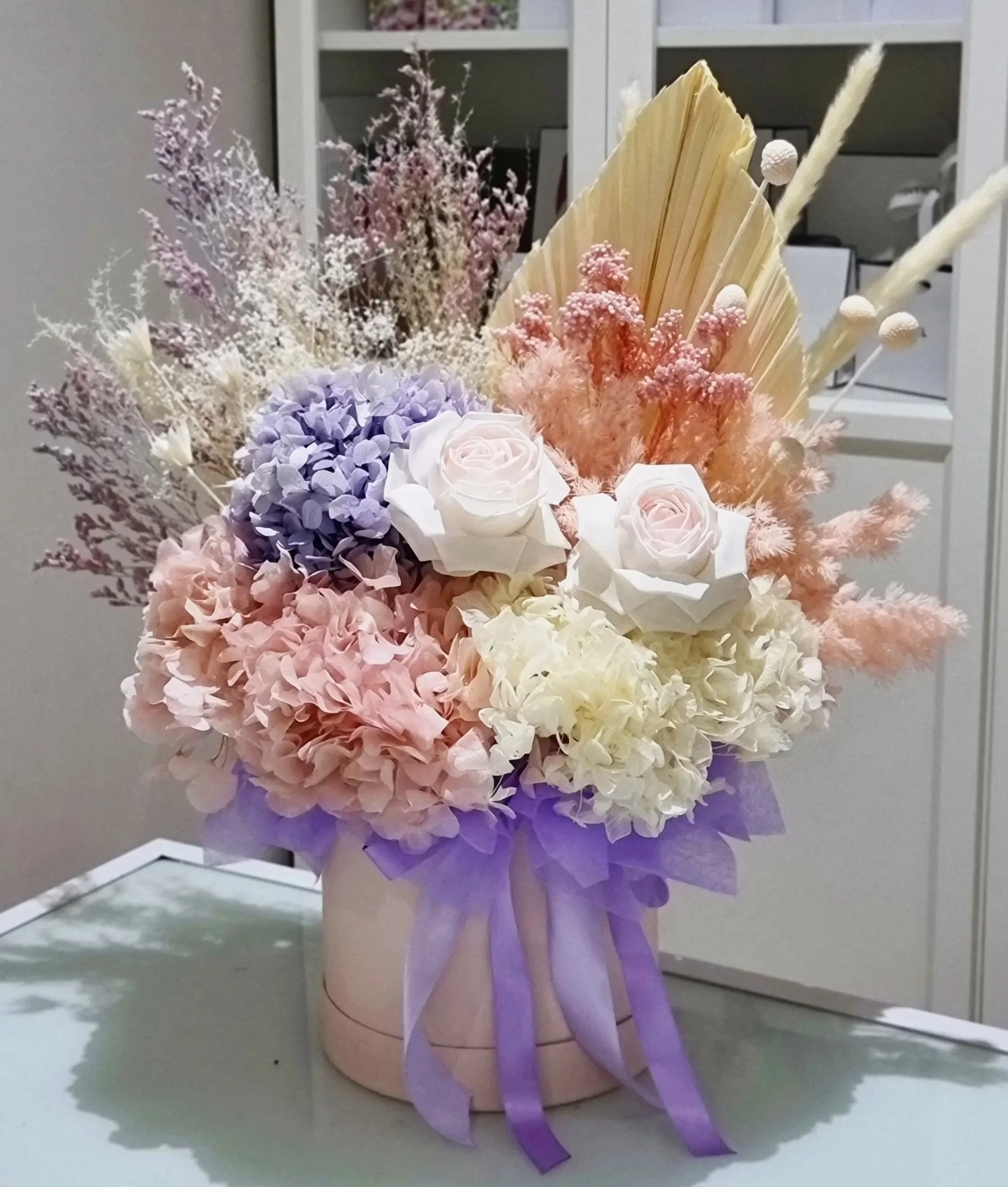 A pastel-coloured flower arrangement in a pink hatbox with purple ribbon, featuring roses, hydrangeas, dried leaves, grasses, and palm leaf.