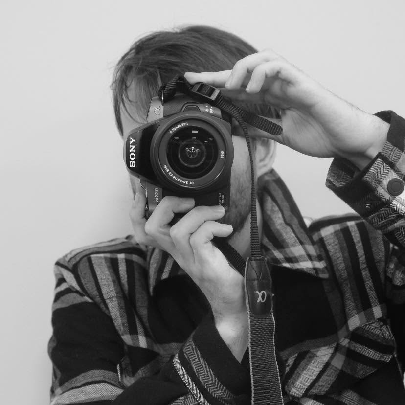A person taking a photo of themselves with a camera, with the camera covering their face. The individual has light-colored hair and is wearing a plaid shirt. The image is in black and white.