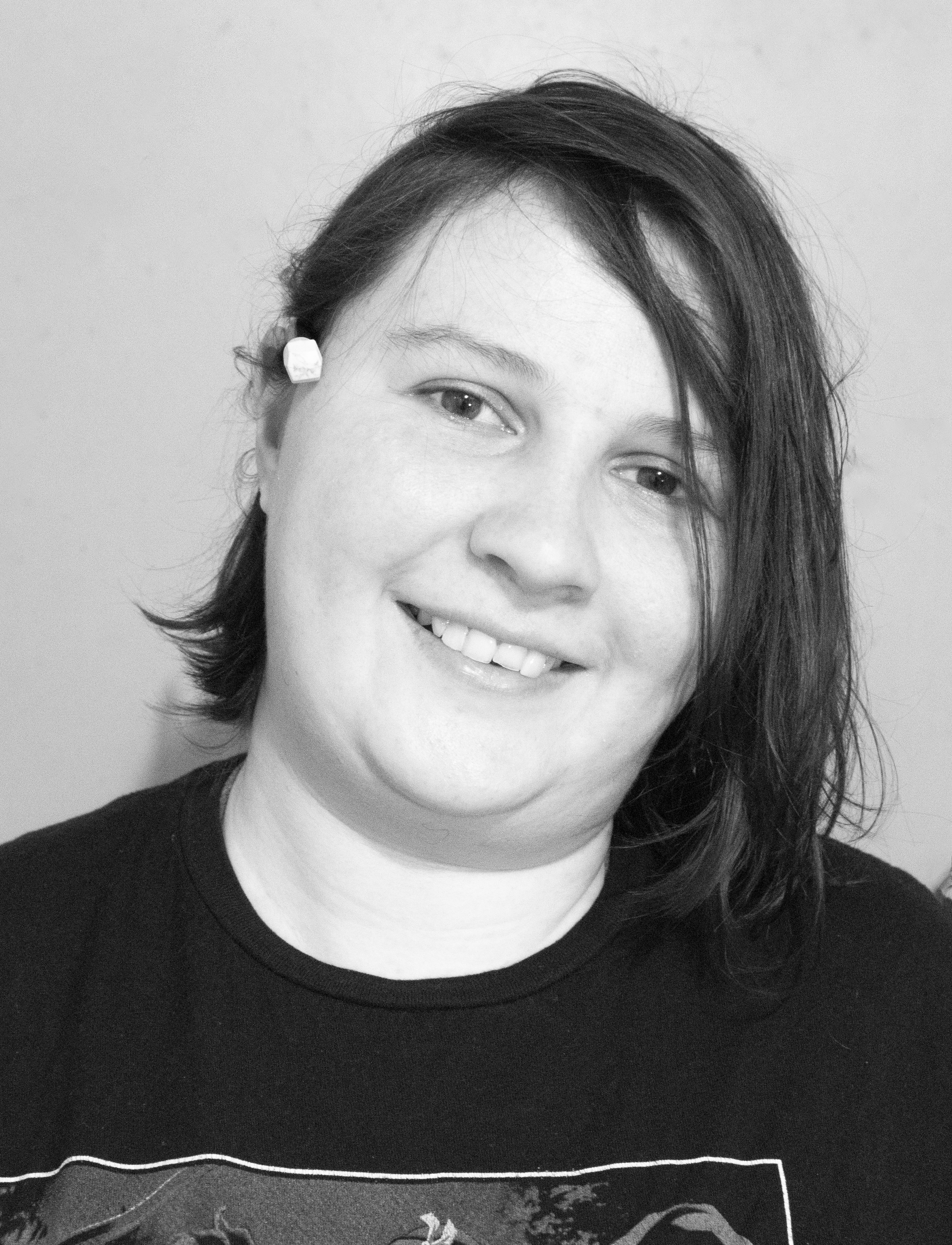 Close-up black-and-white photo of a woman smiling, showing her teeth, with shoulder-length dark hair and wearing a dark shirt against a plain background.