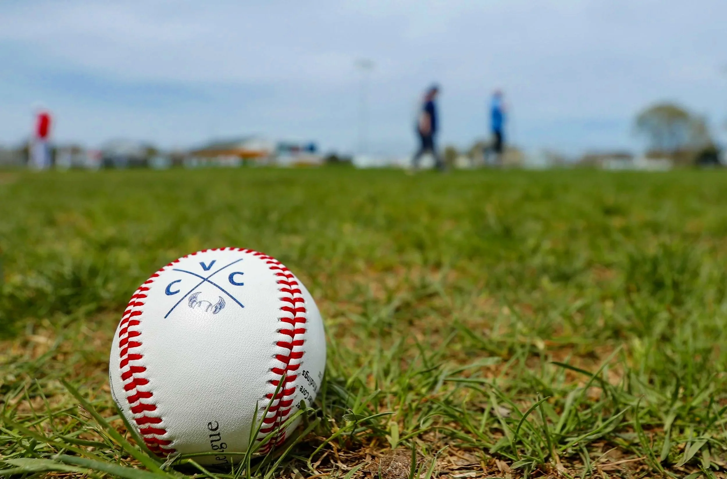 A close-up of a white baseball with red stitching and a blue logo resting on grass, with people playing softball in the blurry background under a cloudy sky.