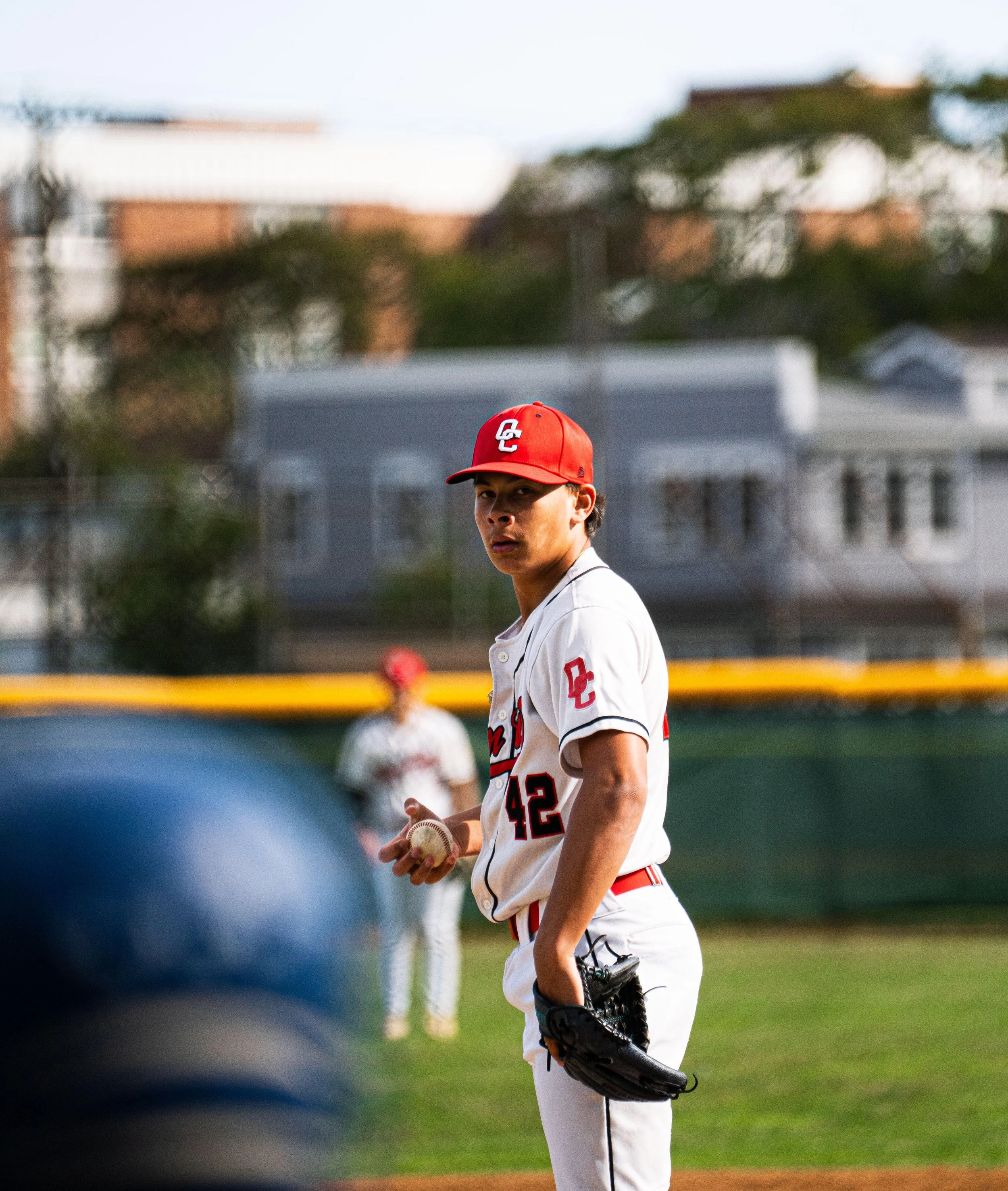 A young baseball player in a white uniform with red accents, wearing a red cap, stands on the field holding a baseball in his right hand and a glove in his left. There is a blurred background with another player and houses.