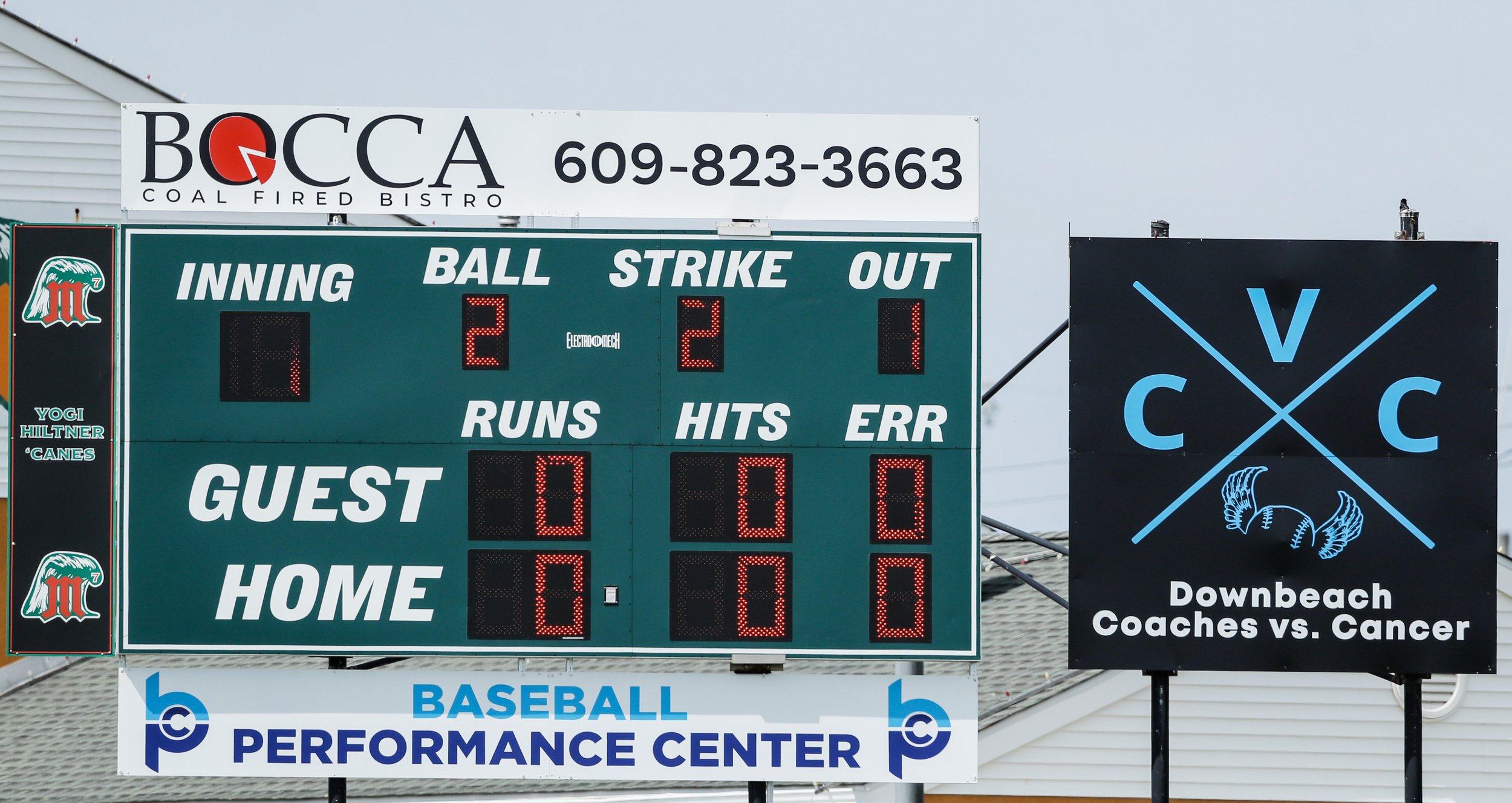 Baseball scoreboard showing the game in the 1st inning with 2 balls, 2 strikes, 1 out, 0 runs, hits, and errors for the guest and home teams. There are advertisements above and below the scoreboard, and a sign about a charity event called 'Downbeach Coaches vs. Cancer' to the right.