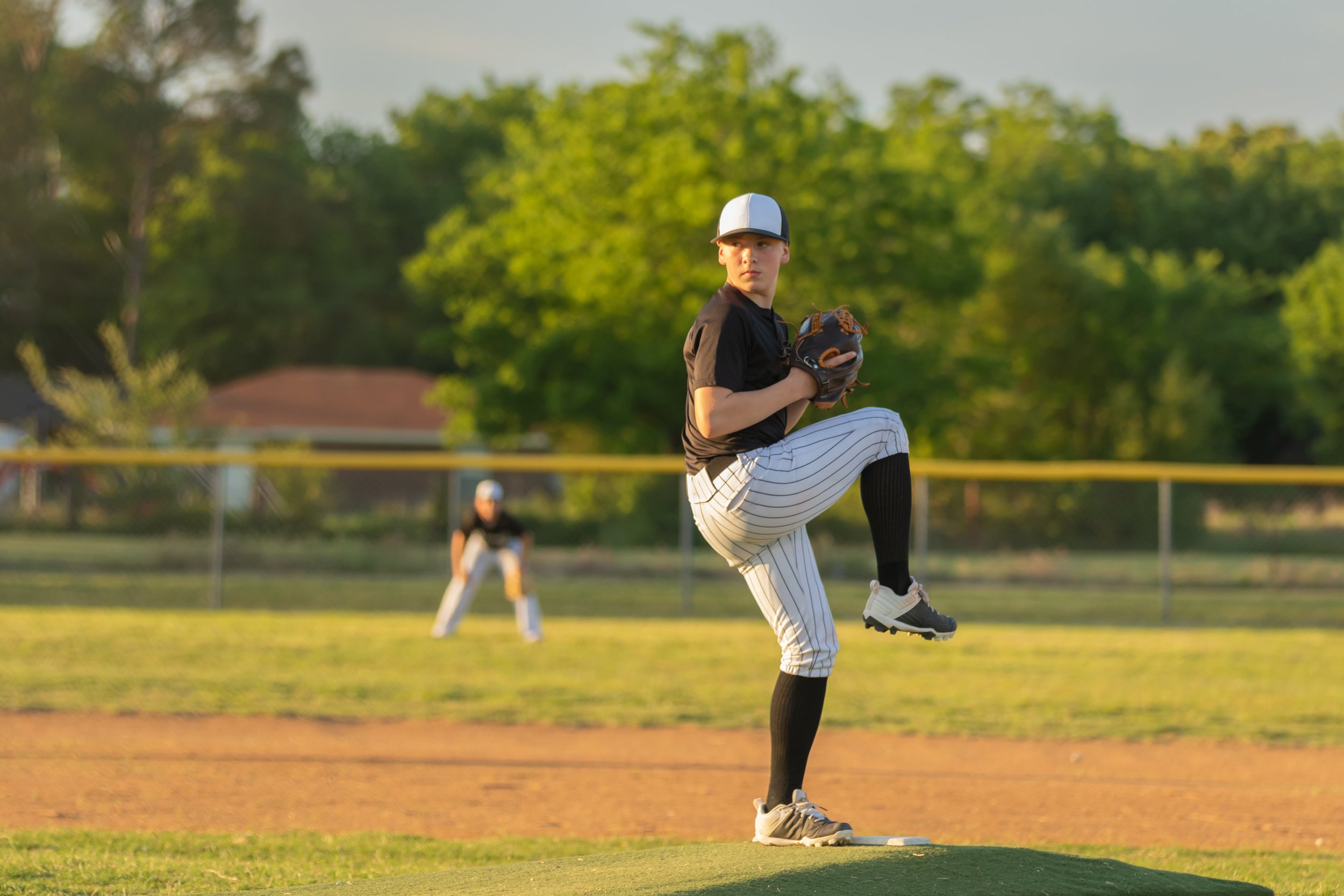 A young baseball player on the field, wearing a black jersey, striped pants, and a white cap, preparing to throw a pitch. In the background, another player is readying to catch, and there are green trees and a fence surrounding the field under a clear sky.