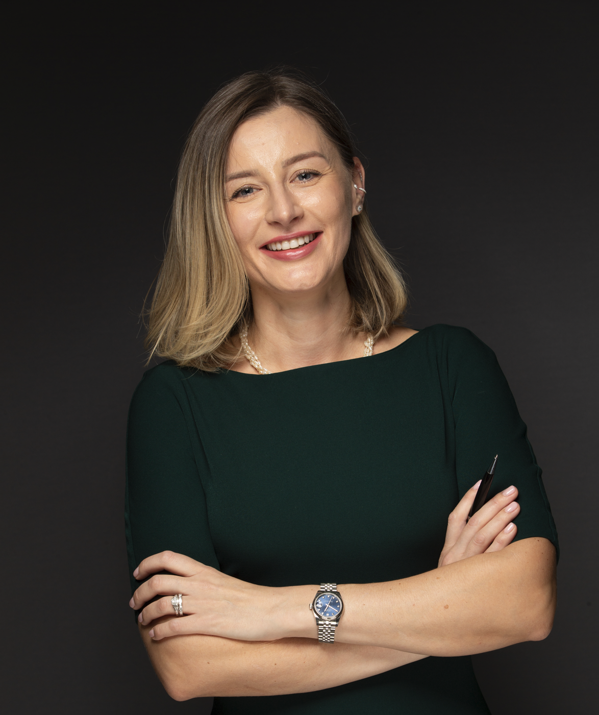 A woman with shoulder-length blonde hair smiling, dressed in a dark green top, standing against a black background, wearing jewelry and a watch.