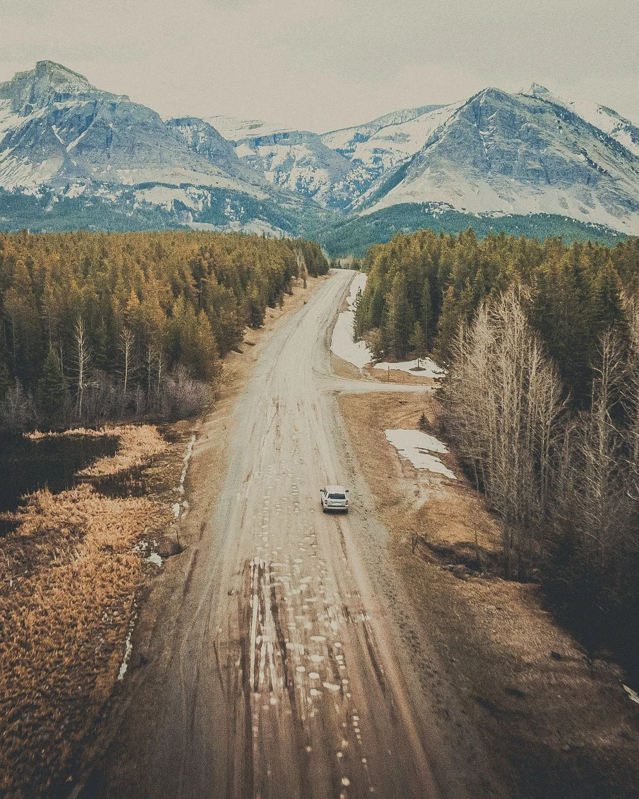 A white car driving on a dirt road in a mountainous area with pine trees and patches of snow, with snow-capped mountains in the background.