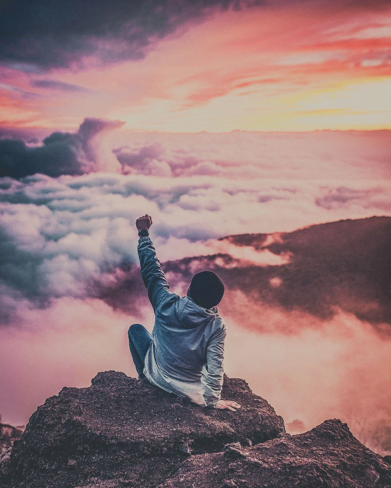 A person sitting on a rocky mountain peak raising one fist in the air, with a view of clouds and a colorful sky during sunset or sunrise.