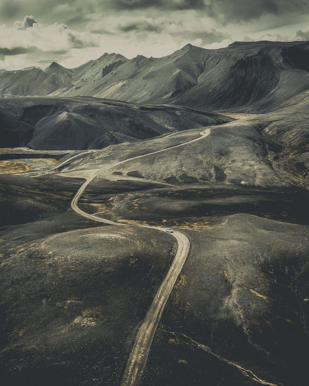 A winding dirt road running through a barren, volcanic landscape with mountains in the background under a cloudy sky.