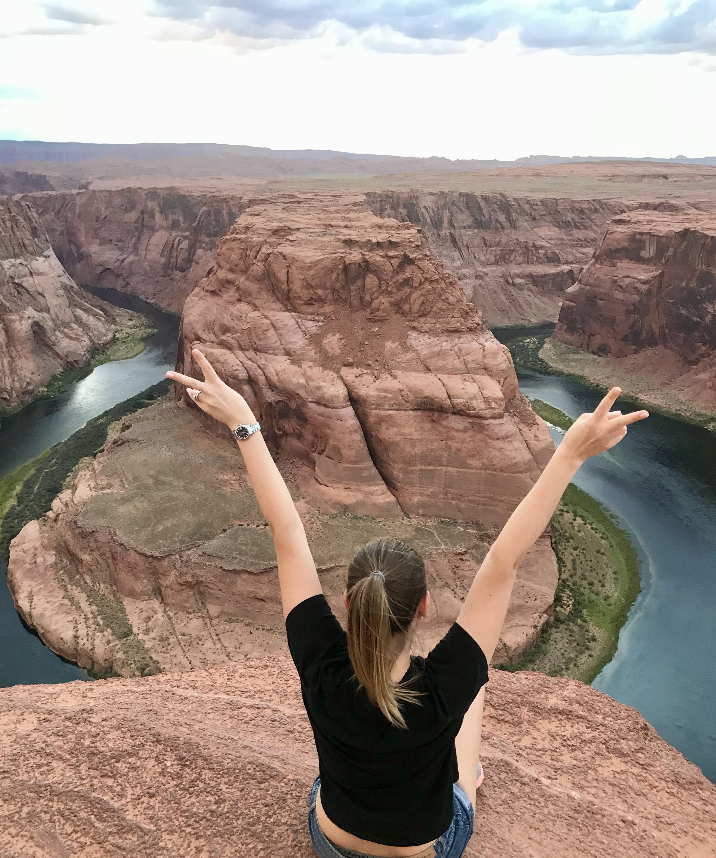 A woman with a ponytail, wearing a black t-shirt and watch, sitting on a cliff and raising her arms with peace signs, overlooking Horseshoe Bend and the Colorado River in a desert landscape.