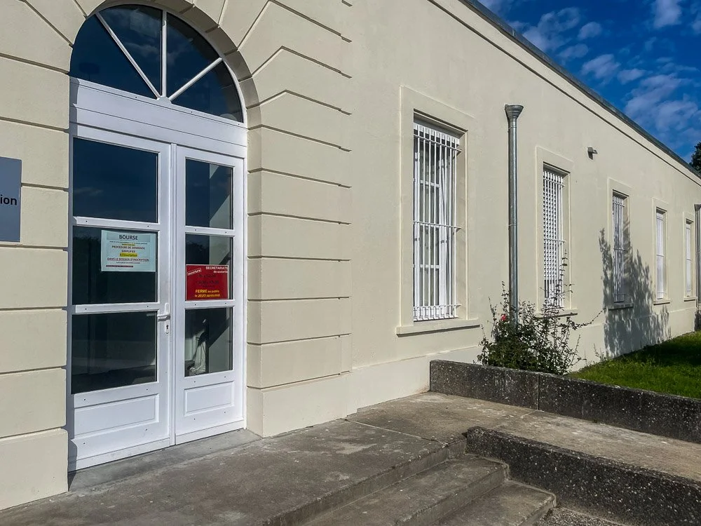 Façade d'un bâtiment toulousain avec une entrée en menuiserie blanche PVC et plusieurs fenêtres avec des grillages blancs, sous un ciel bleu avec quelques nuages.